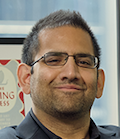 Ibrahim Ahmad, a man of Irani and Pakistani descent, smiles at the viewer. He wears black glasses, a black suit, and has short, black hair.