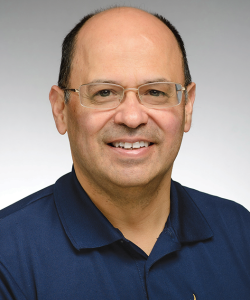 A photo portrait of Francisco Aragón, the director of Letras Latinas. He is a balding middle-aged Latino man with glasses and a blue Letras Latinas polo. He is smiling and standing against a gray background.