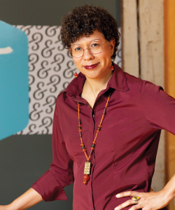 Arleta Little, a Black woman with close-cropped curls and thin-framed glasses, smiles with one hand on her hip. She wears a maroon collared shirt and beaded necklace.