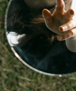 A circular mirror lies flat on the grass. In the reflection, a hand reaches toward the viewer.